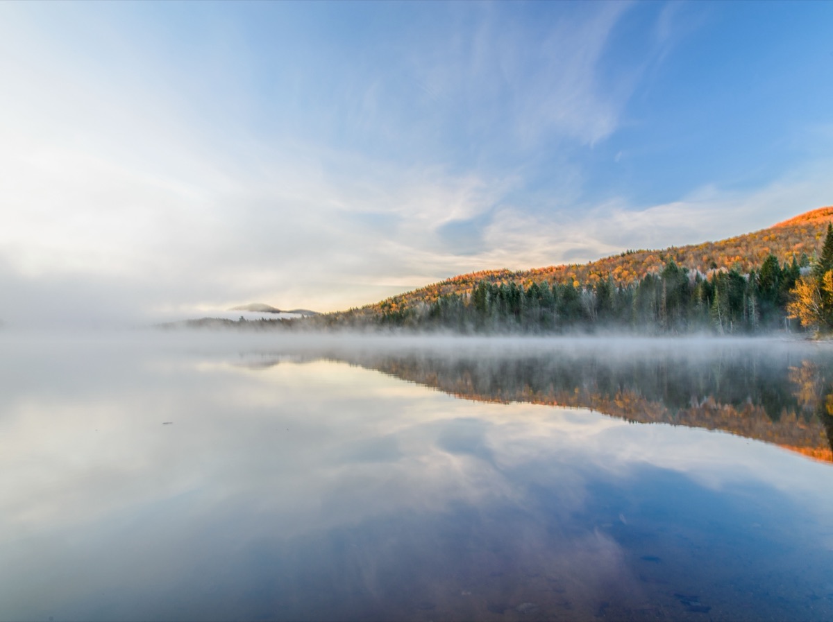 A forested ridge in autumn colour beside a still lake, mist lying on the water under a pale blue sky